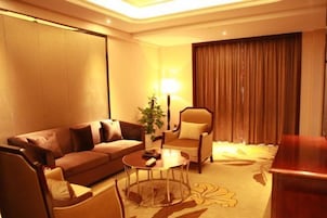 Una habitación de hotel con un sofá marrón, una mesa de centro redonda, una silla, una lámpara y una cortina.