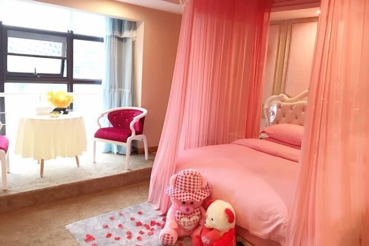 Une chambre à coucher décorée dans des tons roses, avec un lit à baldaquin, une petite table surmontée d’un vase jaune et une chaise rouge.