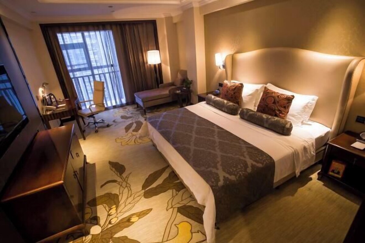 Une chambre d’hôtel avec un grand lit, un bureau, une chaise et une vue sur la ville grâce à de grandes fenêtres.