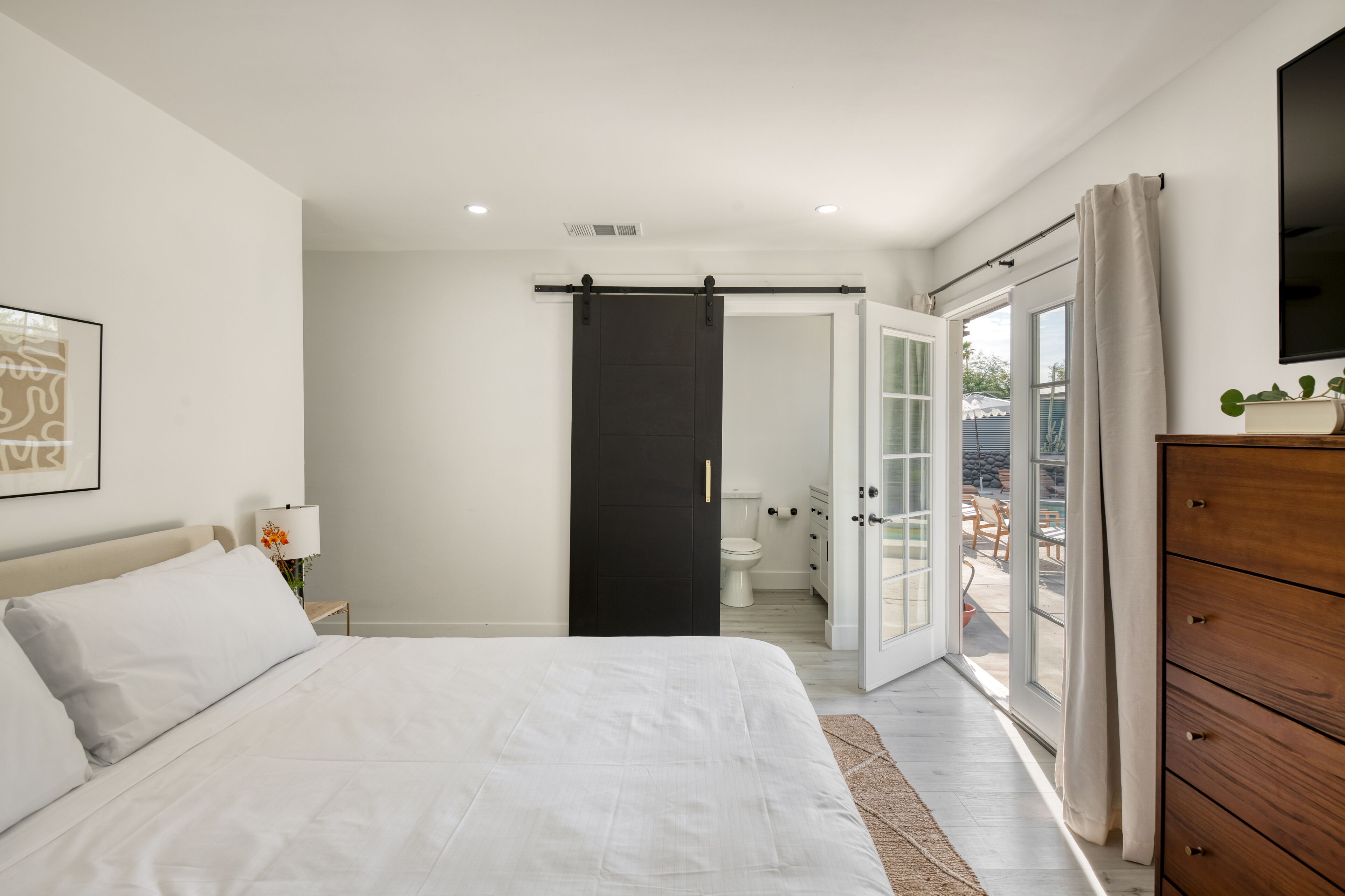 Une chambre à coucher comprenant un lit, une commode en bois, un téléviseur et une porte-fenêtre coulissante donnant sur un espace extérieur.
