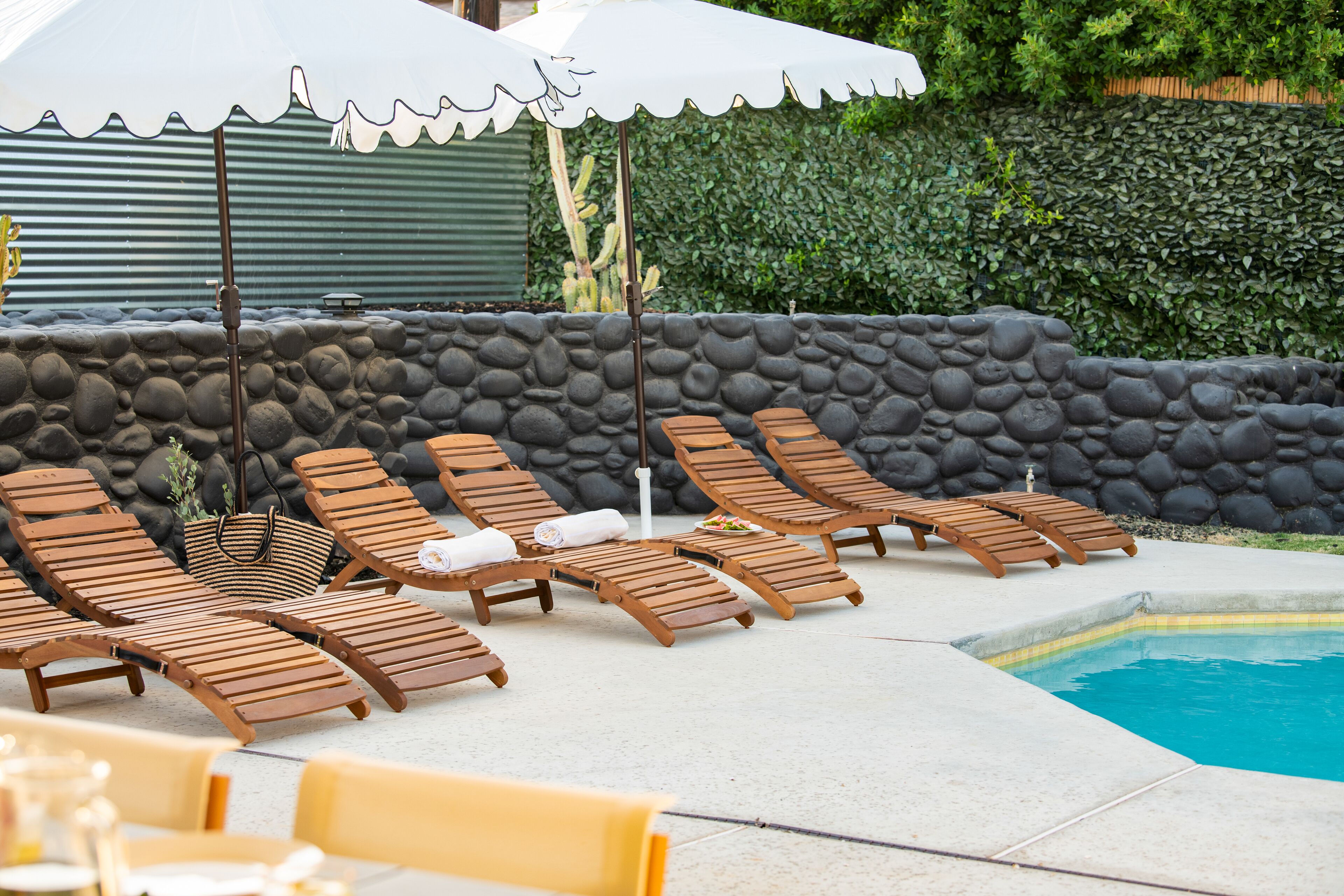 Zona junto a la piscina con sillones de madera, sombrillas, una pared de piedra y una piscina.