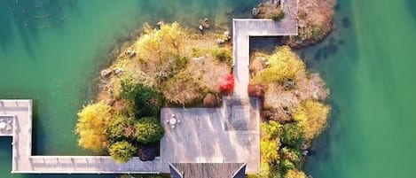 Vue aérienne d’une propriété au bord du lac, avec un quai en bois, un petit pont et un élément décoratif aquatique circulaire.