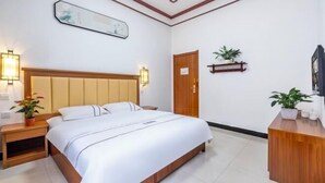 A hotel room with a bed, bedside tables, a wooden headboard, a wall-mounted shelf with plants, a door, and a framed picture on the wall.