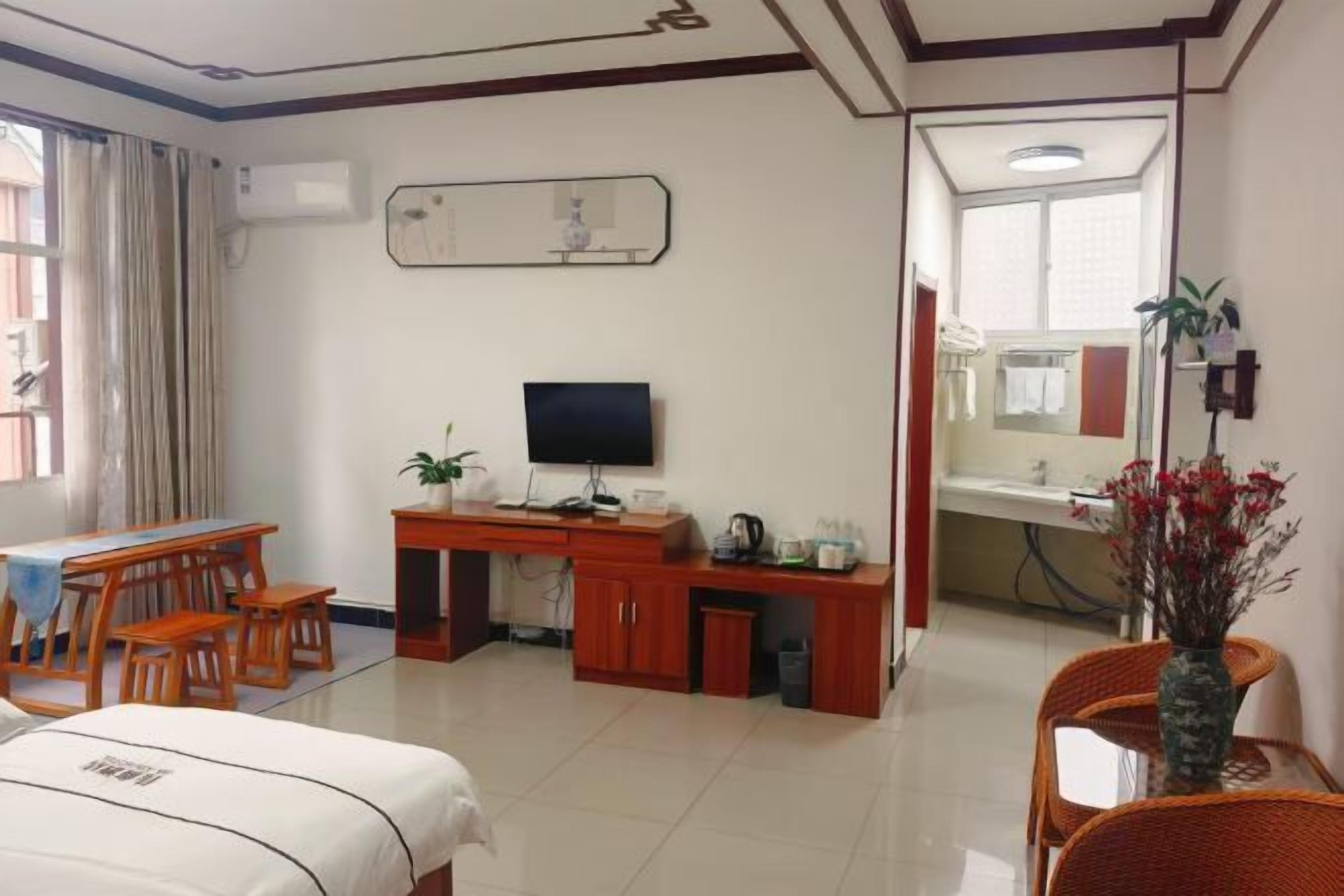 Habitación de hotel con cama, escritorio con computadora, televisor, pequeña mesa con sillas, ventana con cortinas y baño visible a través de una puerta abierta.