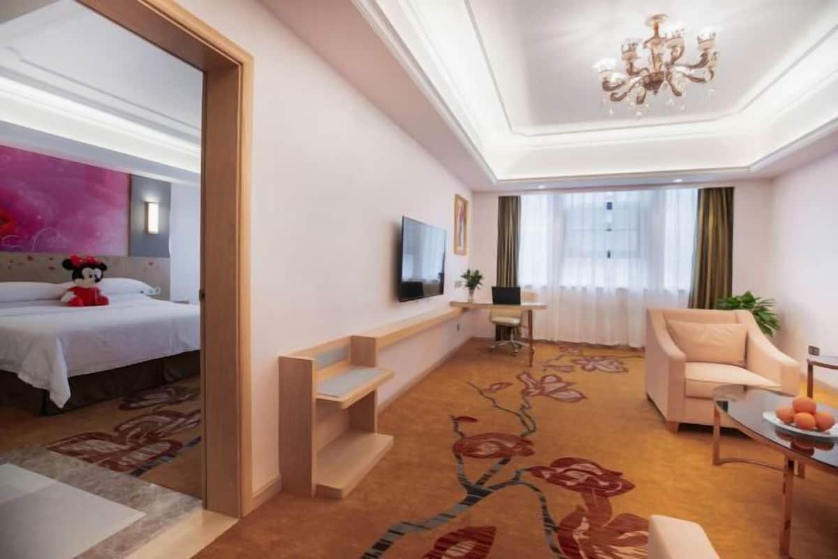 A hotel room with a bed, a desk, a chair, a television, and a chandelier.