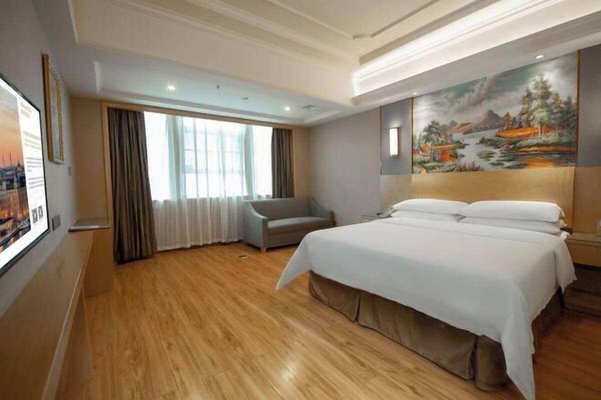 A hotel room with a large bed, a chair, a painting on the wall, and a window with curtains.
