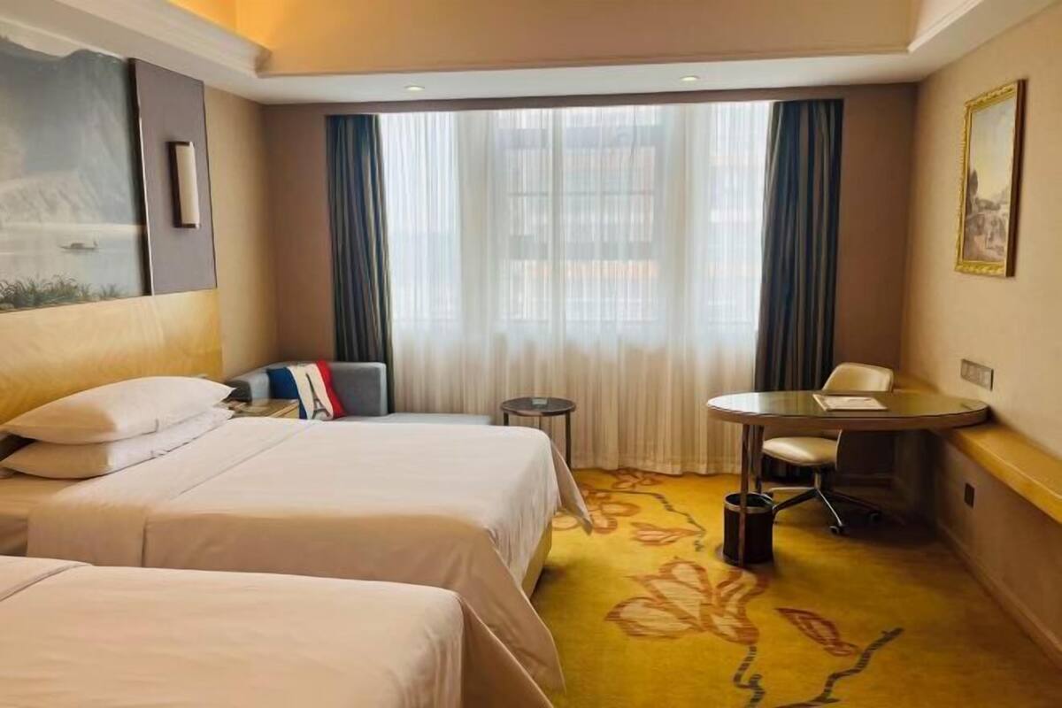 A hotel room with two beds, a small table, a chair, a desk, and a window with curtains.