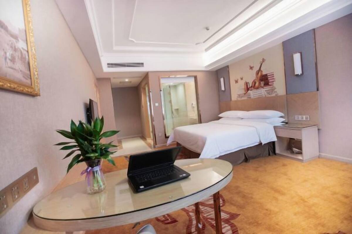A hotel room with a bed, bedside table, and a laptop on a glass table.