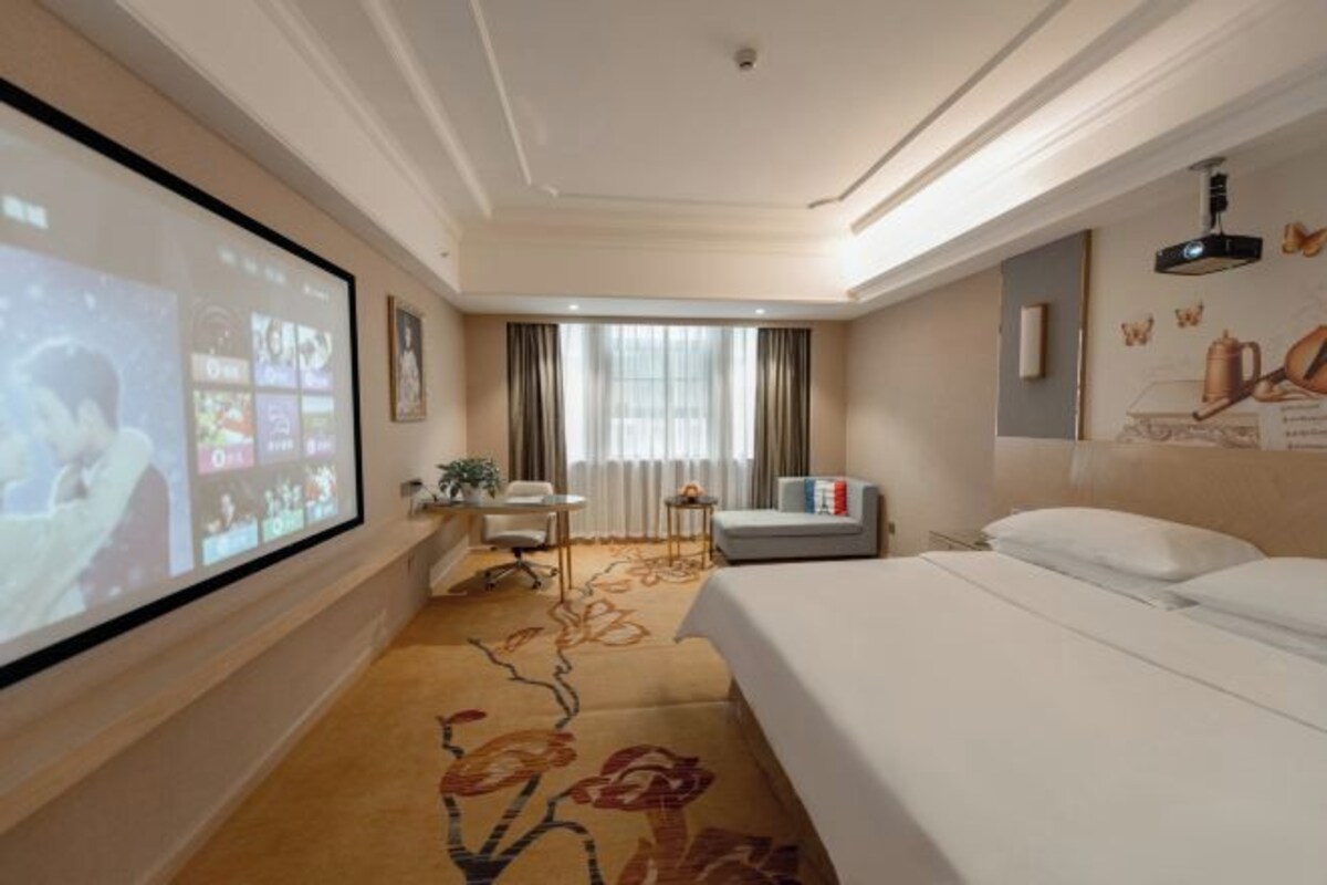 A hotel room with a bed, a desk with a chair, a sofa, a large screen, and a window with curtains.