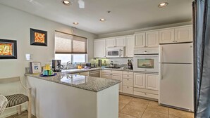 Una cocina moderna con armarios blancos, encimera de granito, microondas integrado, estufa y refrigerador. Hay una ventana con persianas, una silla y una pequeña mesa con una cafetera y tazas.