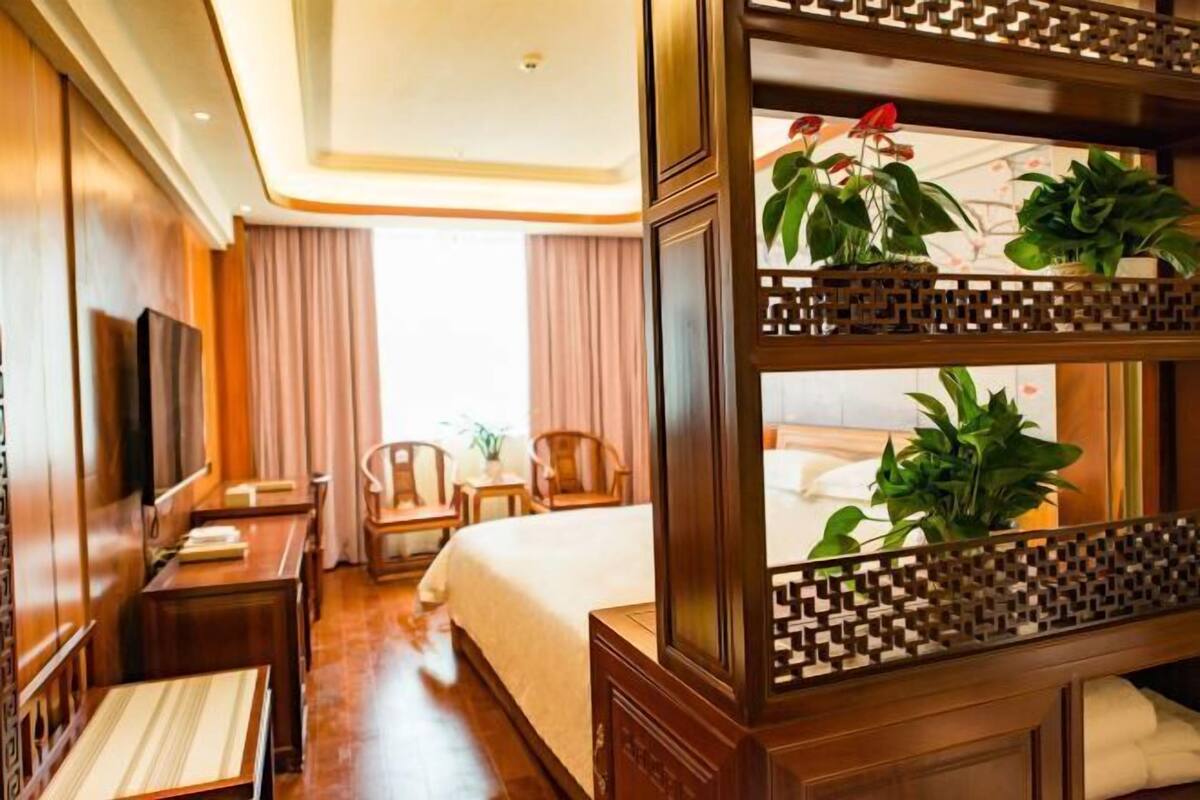A hotel room with a large bed, a TV, a desk, and a chair. There are potted plants on the windowsill.