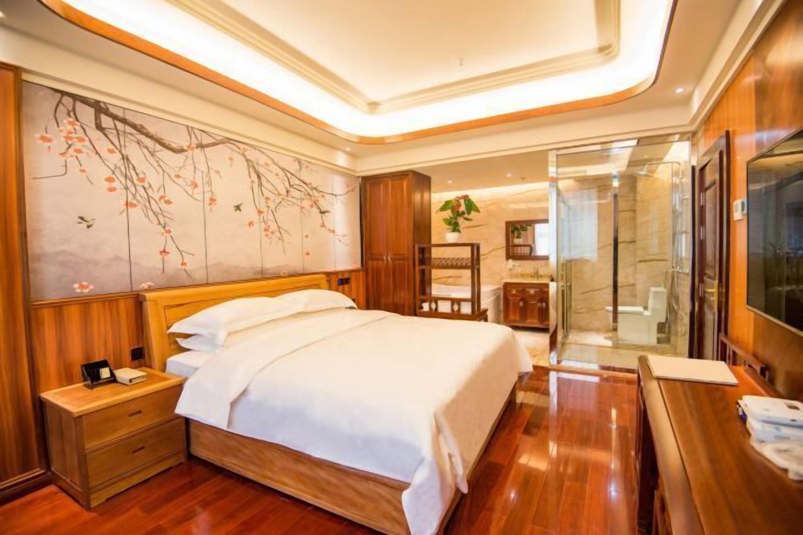 Habitación de hotel con una cama de madera, una mesita de noche, un cuadro grande en la pared, un baño con cabina de ducha de vidrio y un televisor sobre una mesa de madera.