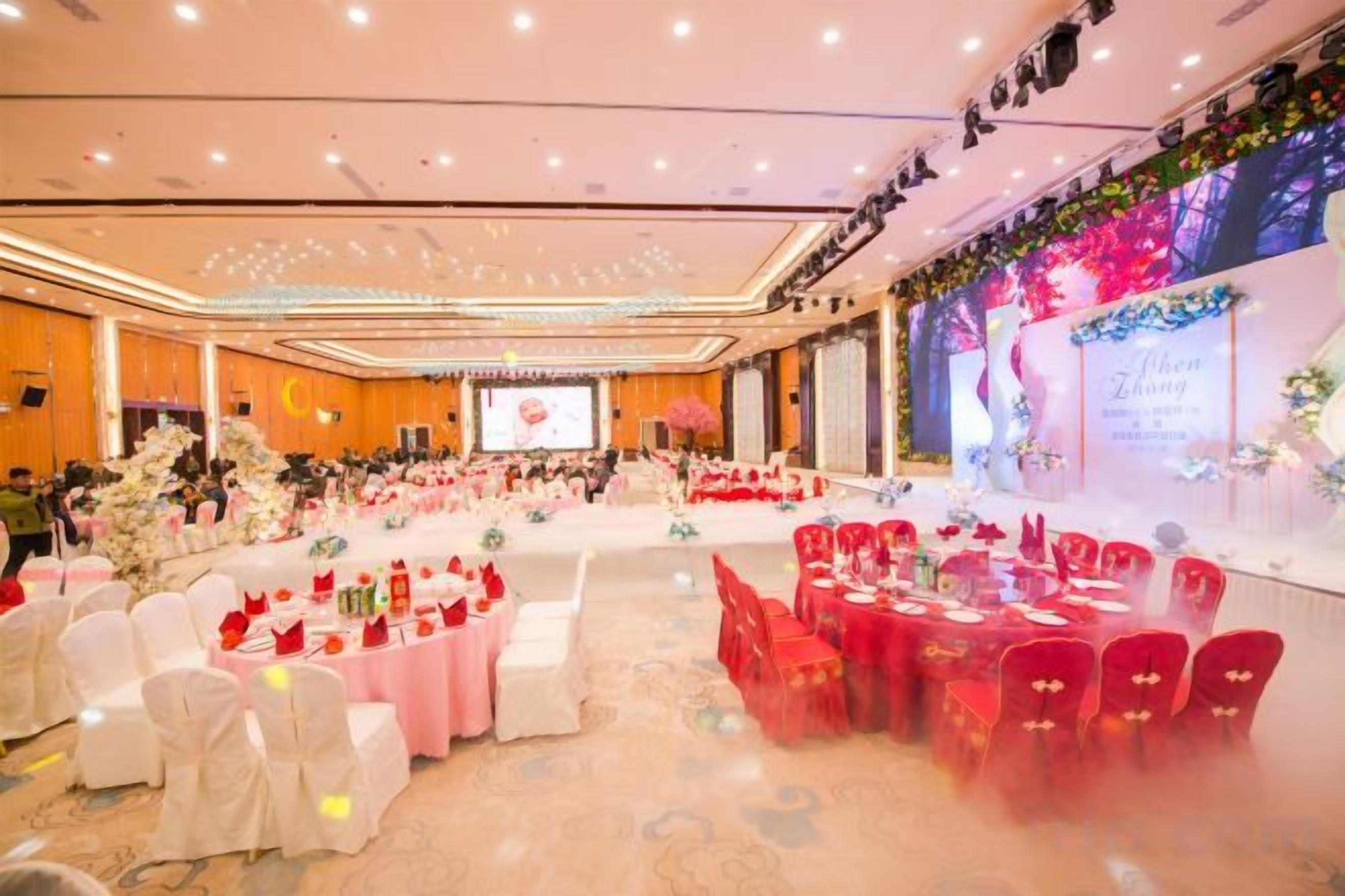 A banquet hall with round tables and chairs, a stage with a screen, and floral decorations.