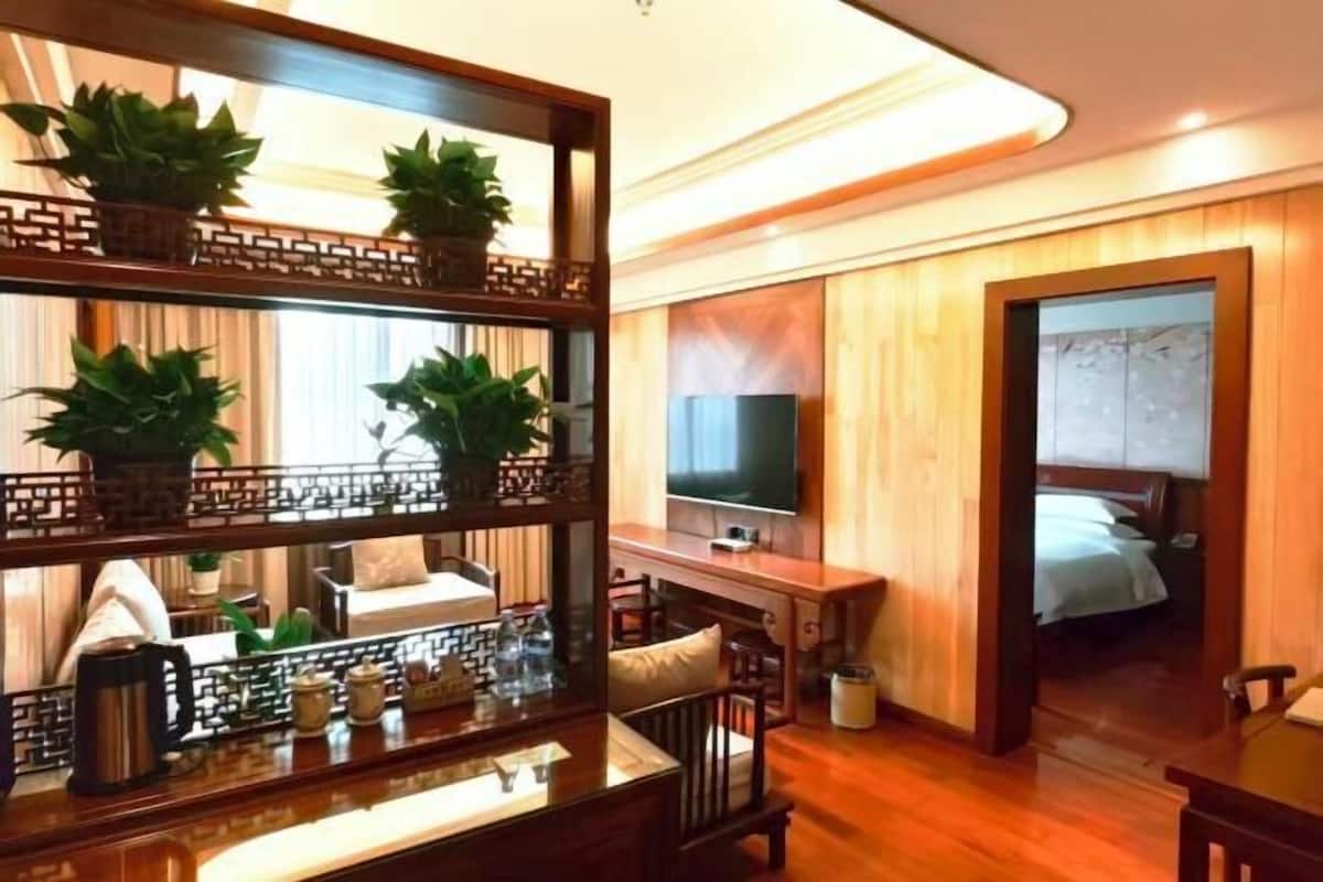 A hotel room with a wooden interior, a TV mounted on the wall, a bed with a white duvet, and a small table with a plant and a teapot.