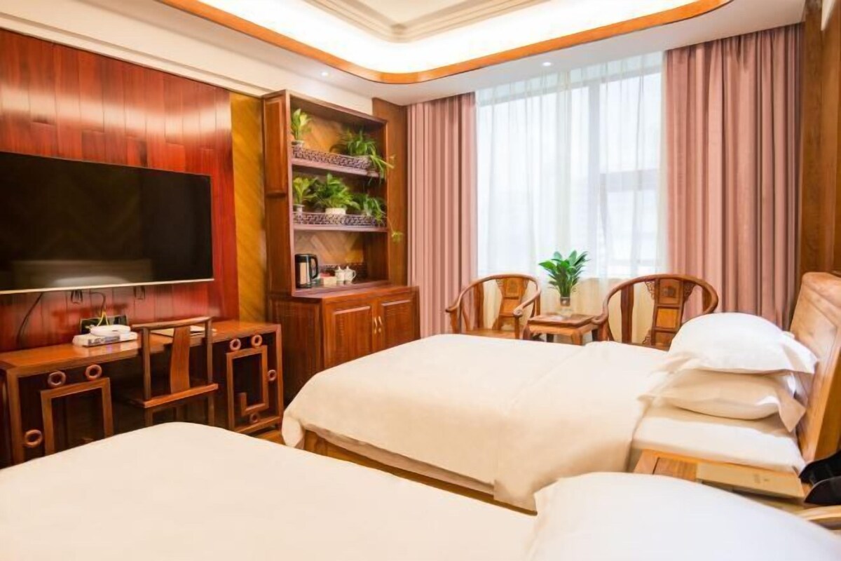 A hotel room with a large flat-screen TV, two beds, a wooden cabinet with plants, and a window with curtains.