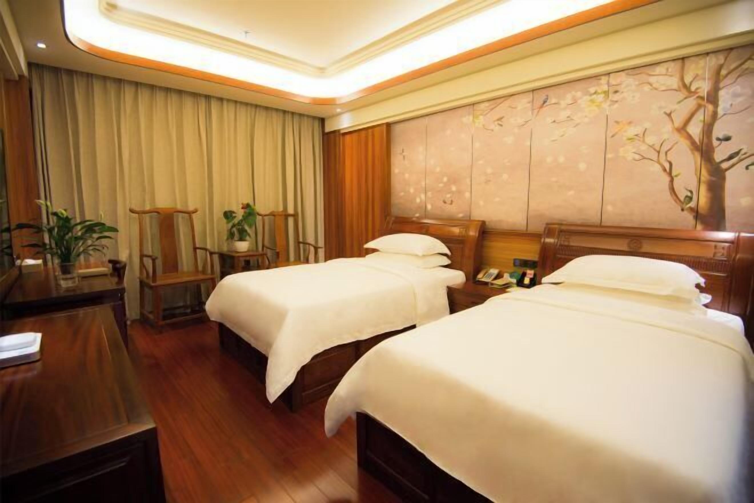 A hotel room with two beds, a desk, a chair, and a wall with a tree mural.