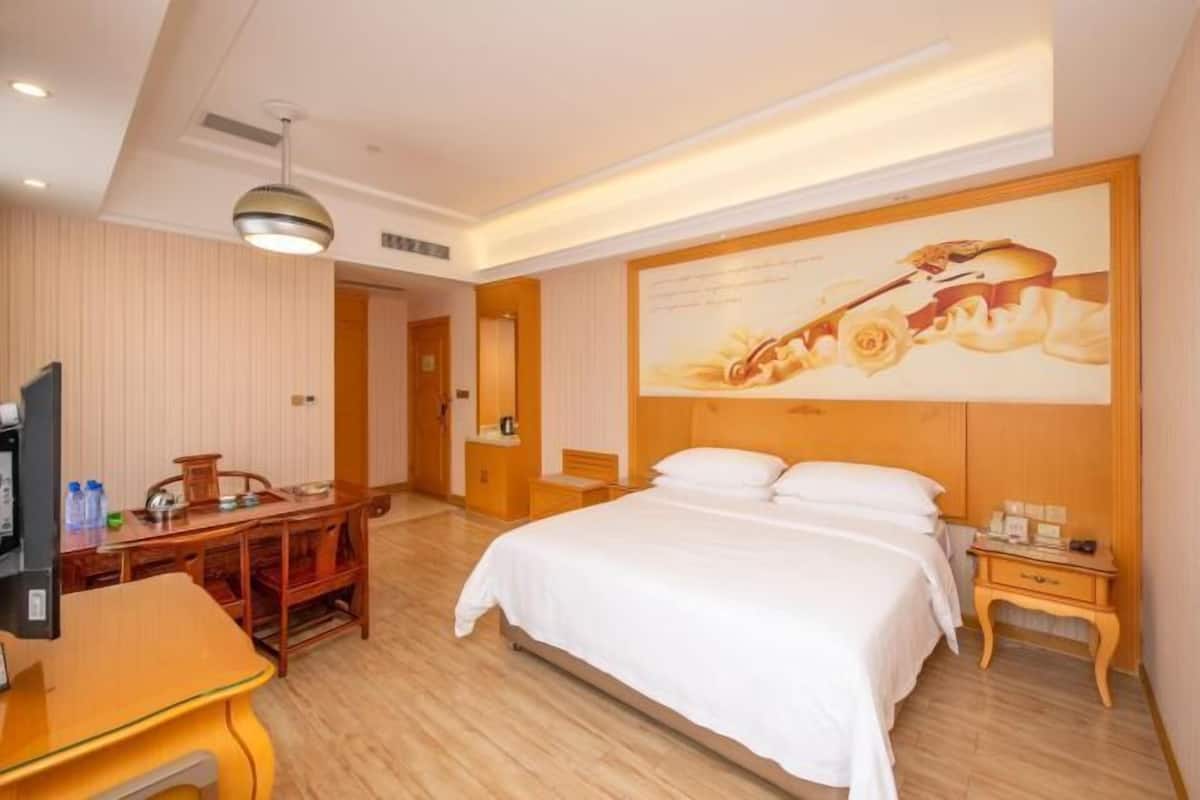A hotel room with a wooden floor, a bed with white linens, a desk with a chair, a television, and a painting on the wall.