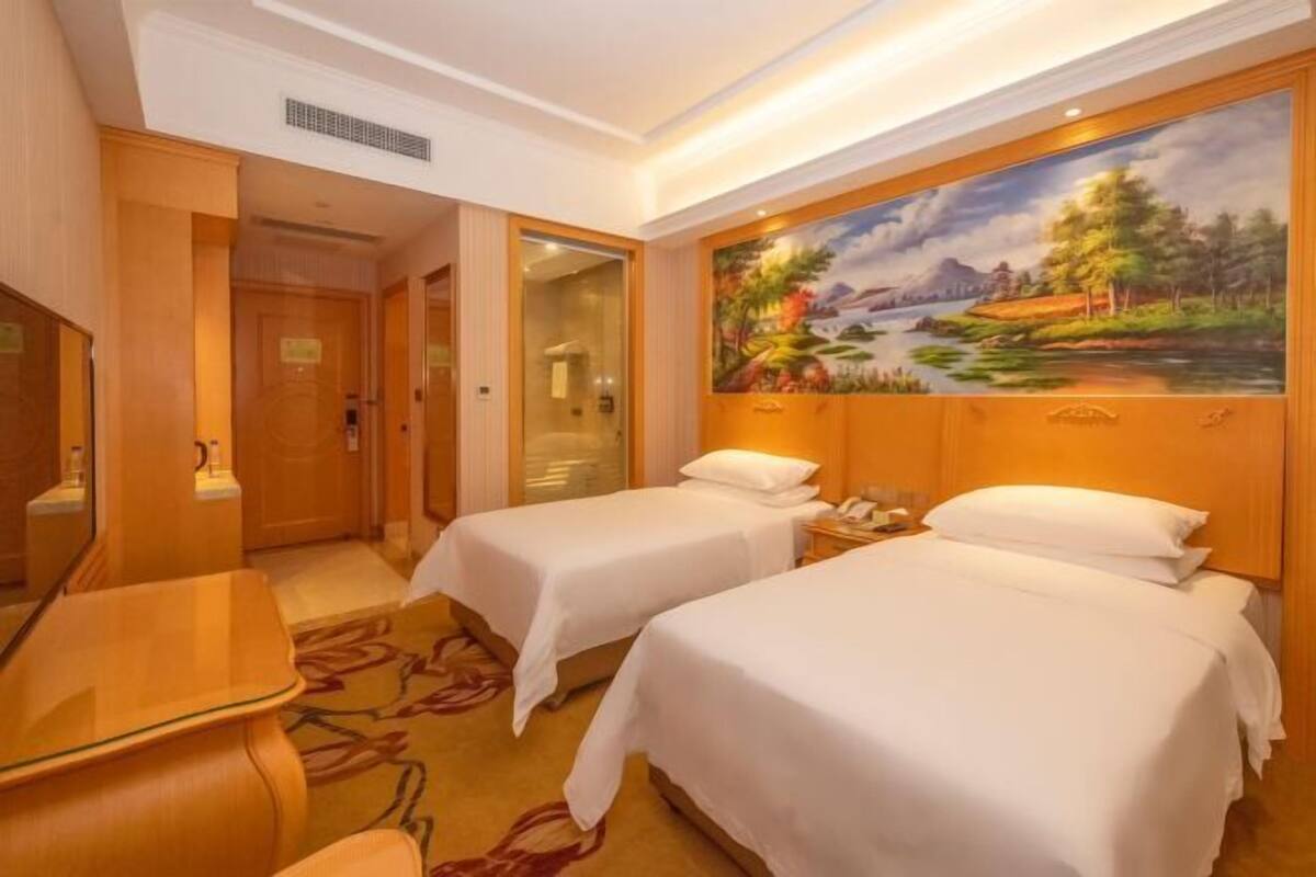 A hotel room with two beds, a desk, and a painting of a landscape.