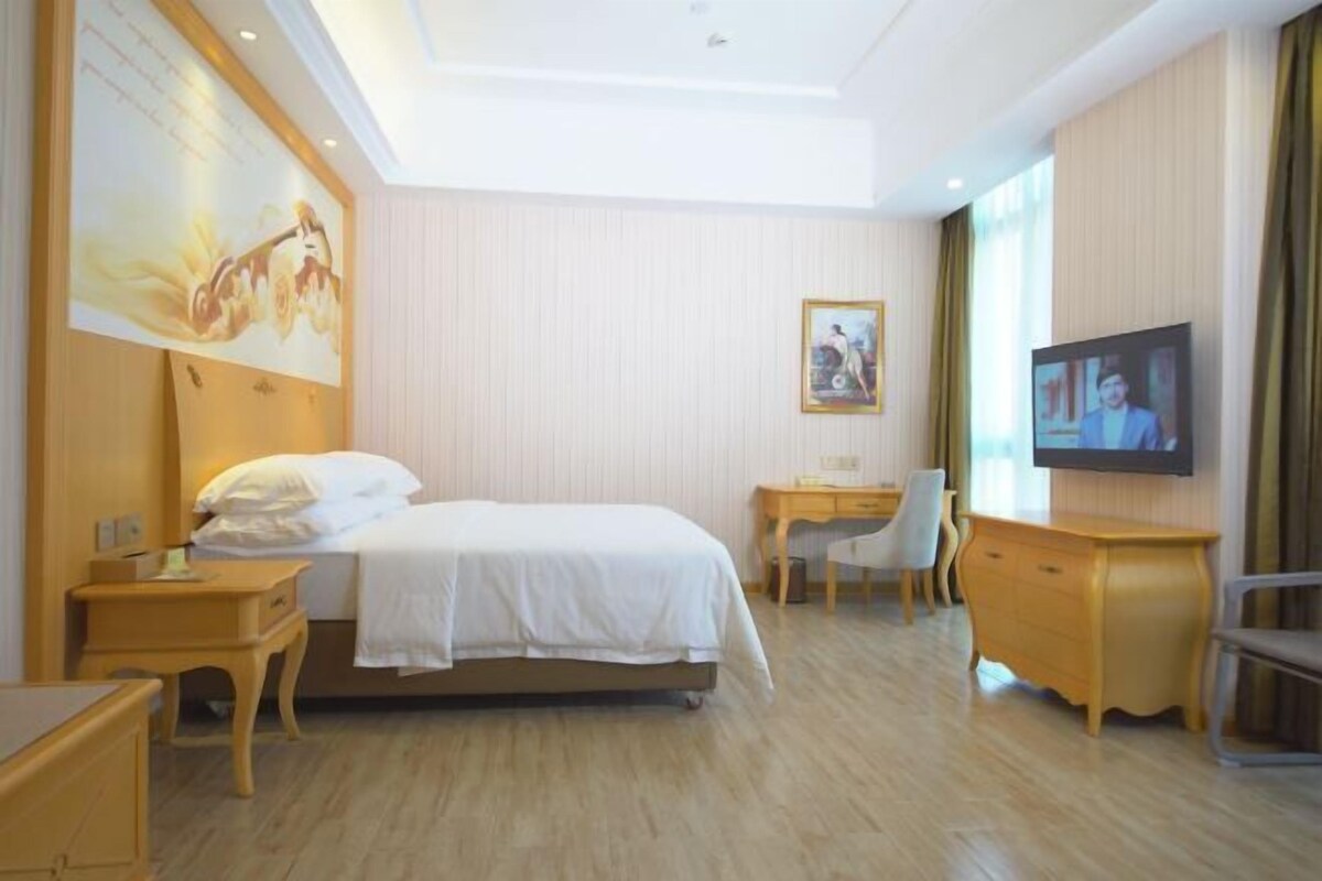 Quarto de hotel com cama, escrivaninha, cadeira, televisão e cômoda.