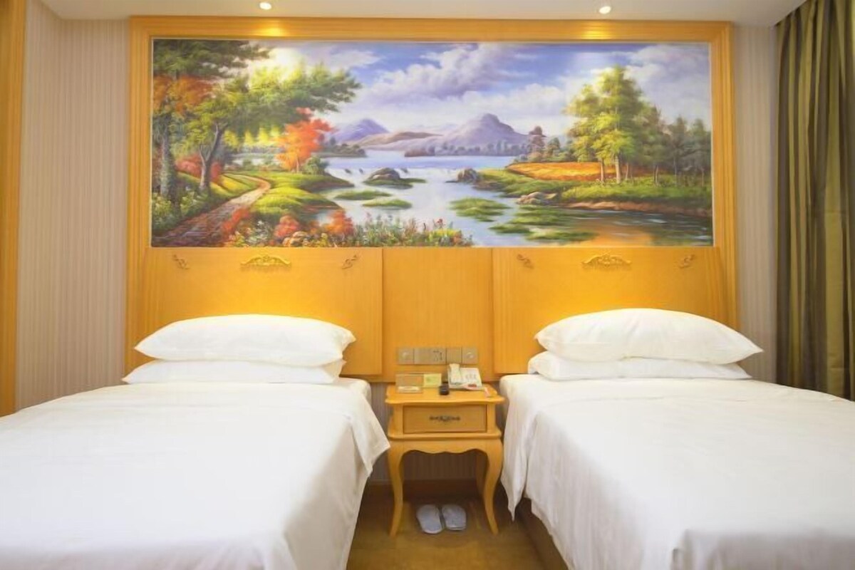 Quarto de hotel com duas camas, um quadro de paisagem na parede e uma pequena mesa de cabeceira de madeira com um telefone.