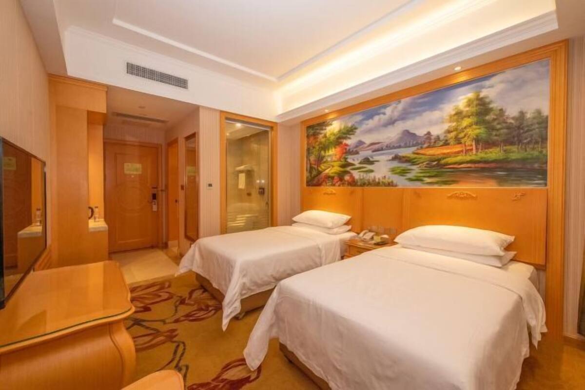 A hotel room with two beds, a painting of a landscape, a desk, and a mirror.