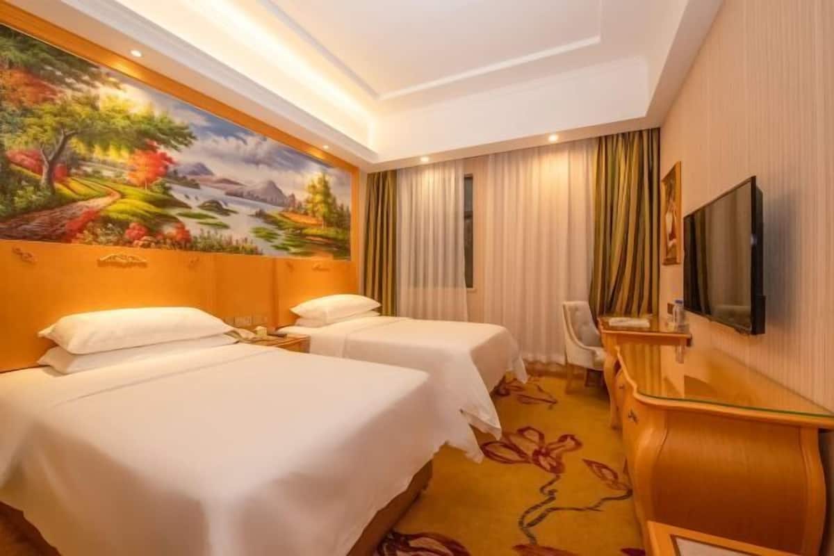 A hotel room with two beds, a desk, a chair, a TV, and a mural on the wall.