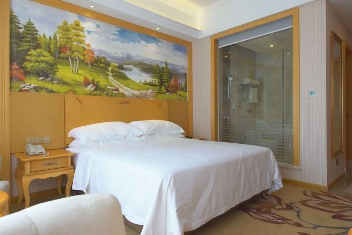 Quarto de hotel com uma cama grande, mesa de cabeceira com telefone, pintura de paisagem na parede e porta de vidro deslizante.