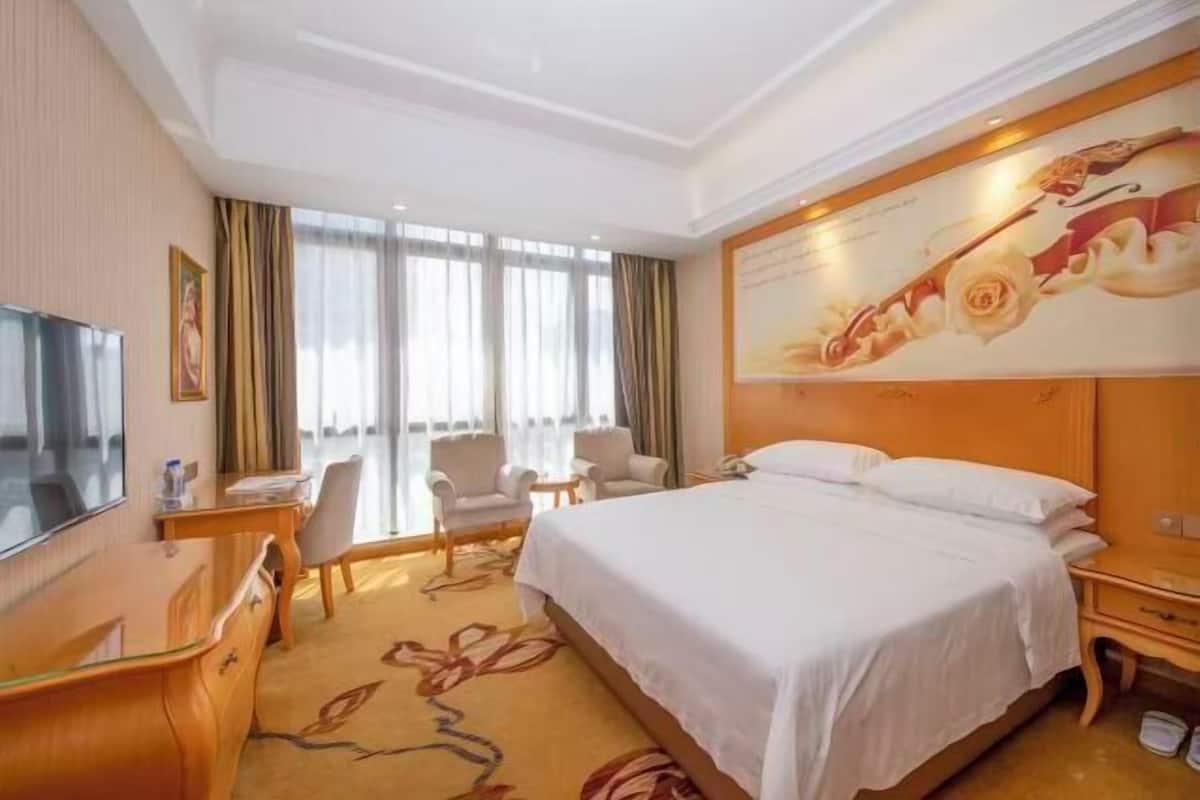 A hotel room with a bed, a desk, a television, and a large window with curtains.