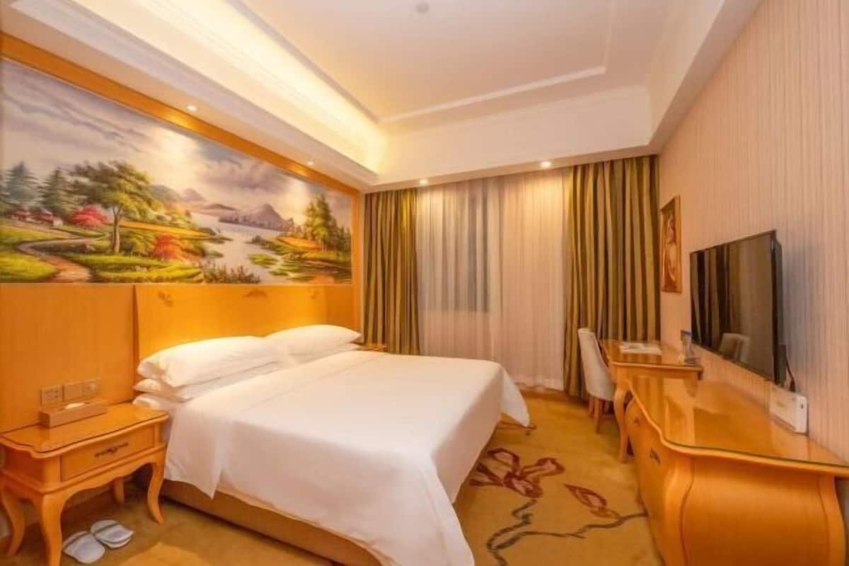 A hotel room with a bed, a desk, a TV, and a painting on the wall.