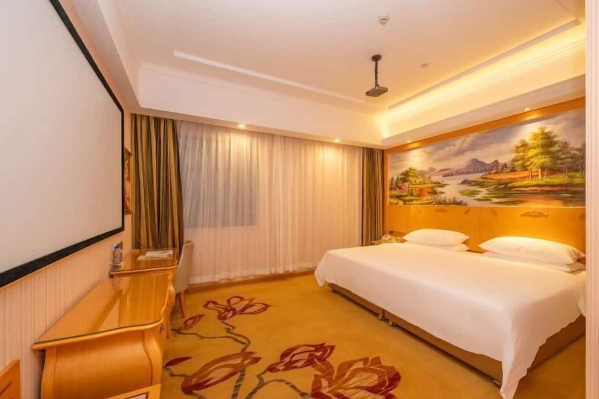 A hotel room with a bed, a desk, a chair, a painting on the wall, and a carpet with floral patterns.