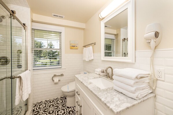 A bathroom with a glass shower enclosure, a sink with a marble countertop, a hairdryer mounted on the wall, and a toilet. - Waterfront Cape May canal property 5min to beach landscaped grounds (Cape May)