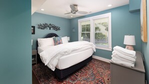 Une chambre à coucher avec un lit, une fenêtre avec des stores, un ventilateur de plafond et une table de chevet avec une lampe.