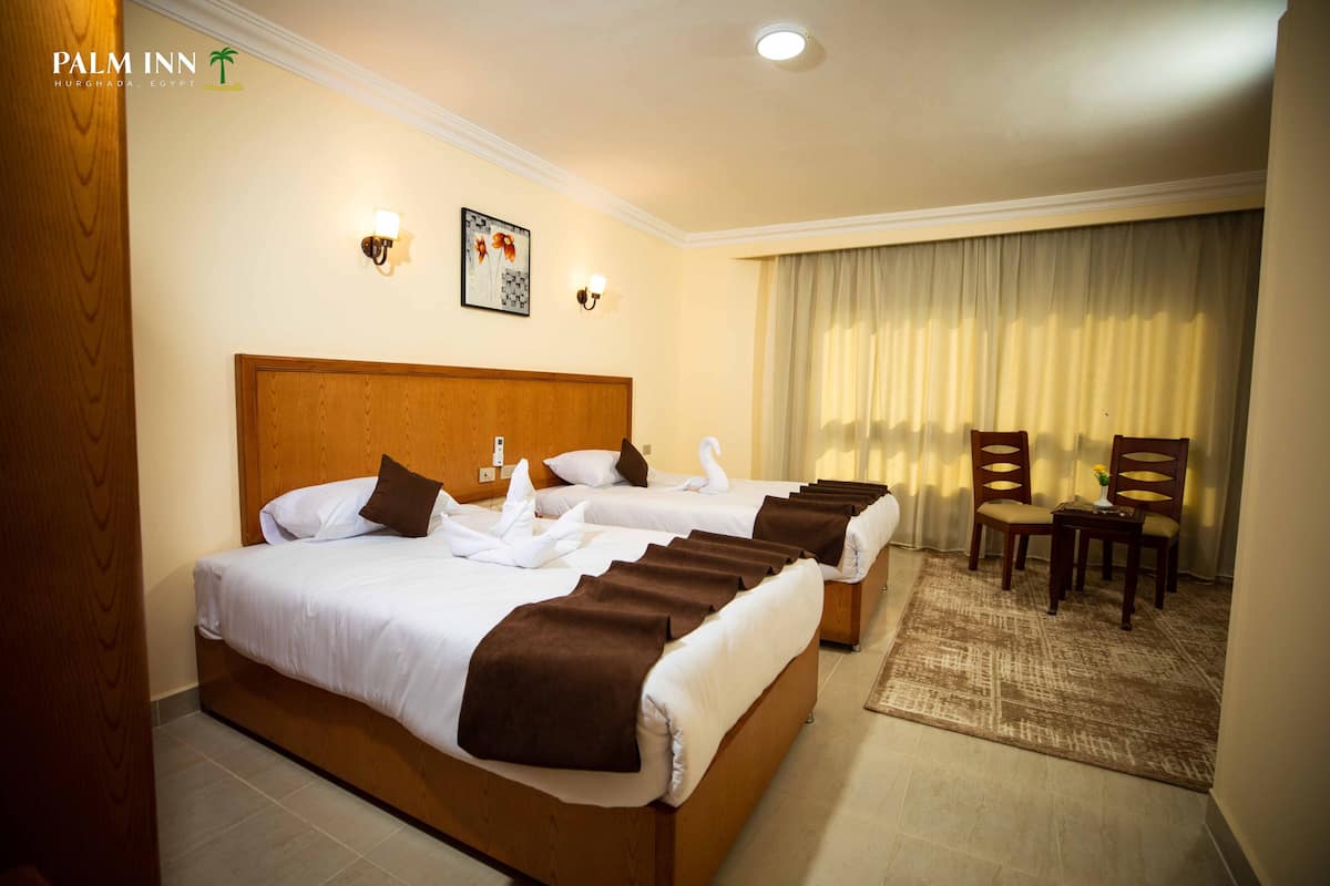 A hotel room with two beds, a wooden headboard, a small table with chairs, and a large window with curtains.