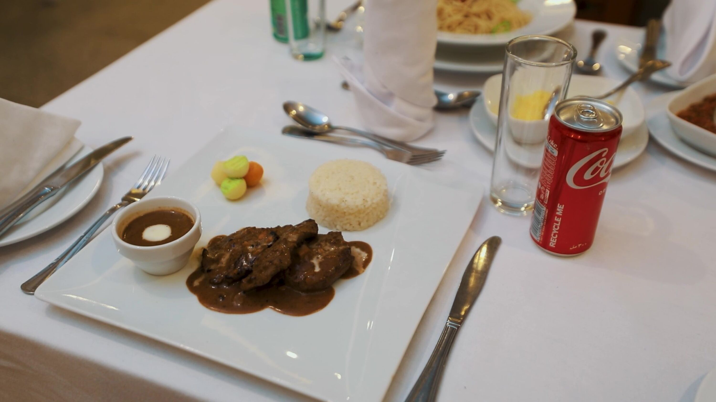 A dining table with a plate of food, utensils, a glass of water, and a can of soda.