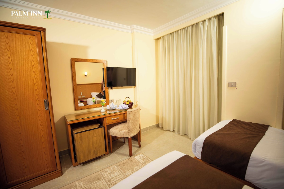 Une chambre d’hôtel avec un bureau en bois, une chaise, un lit recouvert d’une couverture marron, un téléviseur fixé au mur et une grande fenêtre avec des rideaux.