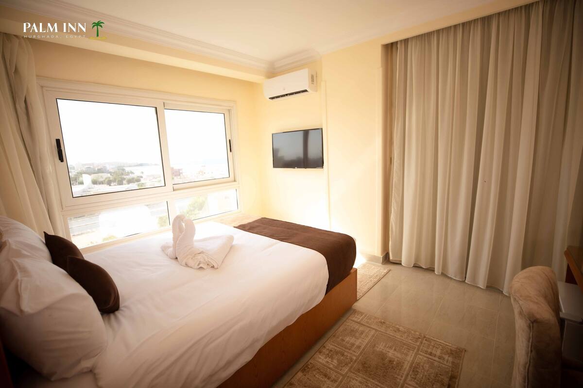 A hotel room with a large bed, a flat-screen TV mounted on the wall, and a window with curtains.