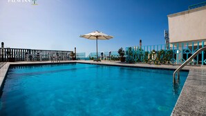 Piscina de color blau clar amb un parasol blanc, envoltada per una tanca blava i un edifici amb el logotip d'una palmera.