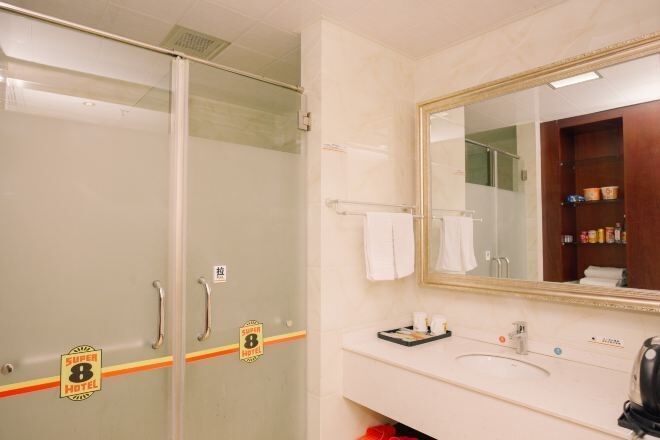 Un baño de hotel con cabina de ducha de vidrio, un lavamanos con artículos de aseo y un espejo con toallero.
