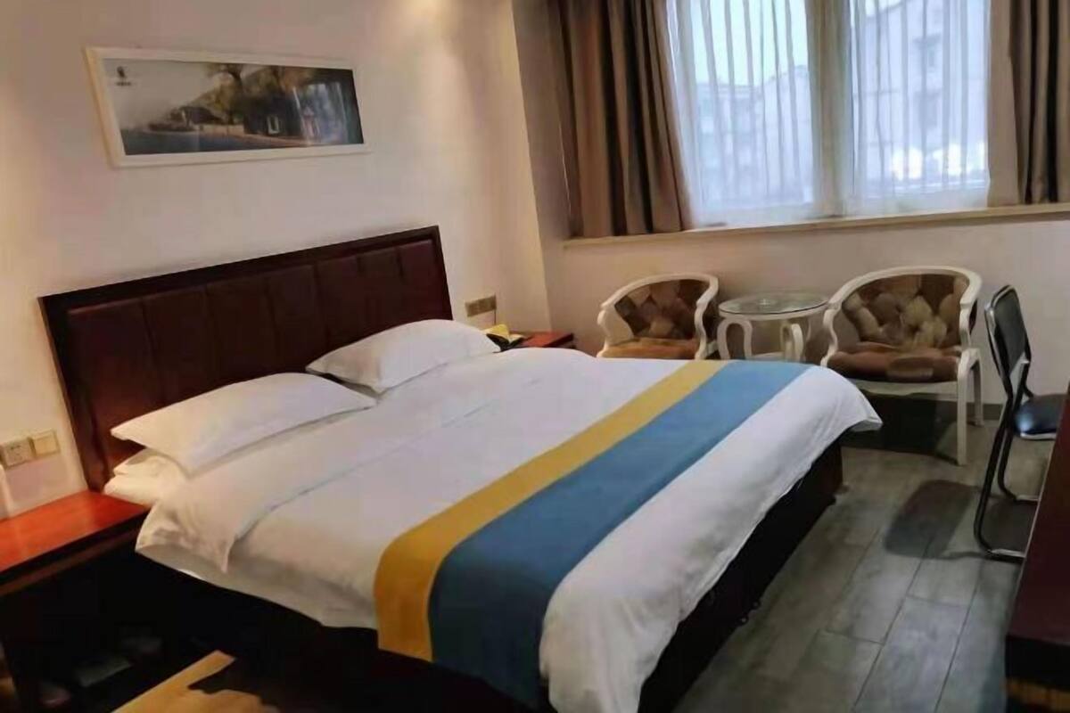 Een hotelkamer met een bed, nachtkastje, stoel, klein tafeltje en een raam met gordijnen.