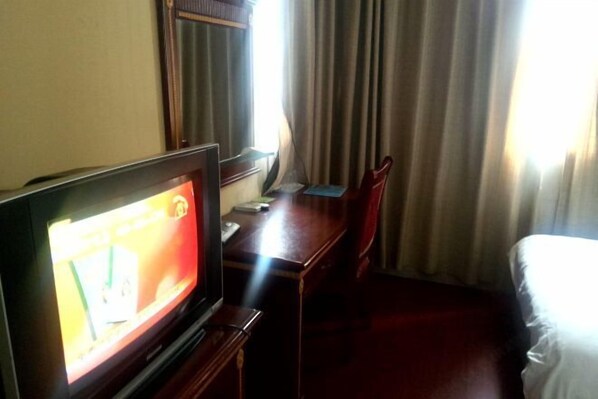 A hotel room with a television on a wooden stand, a desk with a chair, a bed, and a window with curtains. - Haitian Hotel (Wuhu)