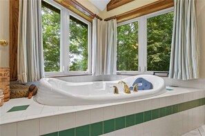 A bathroom with a large bathtub, a window with curtains, and a view of trees.