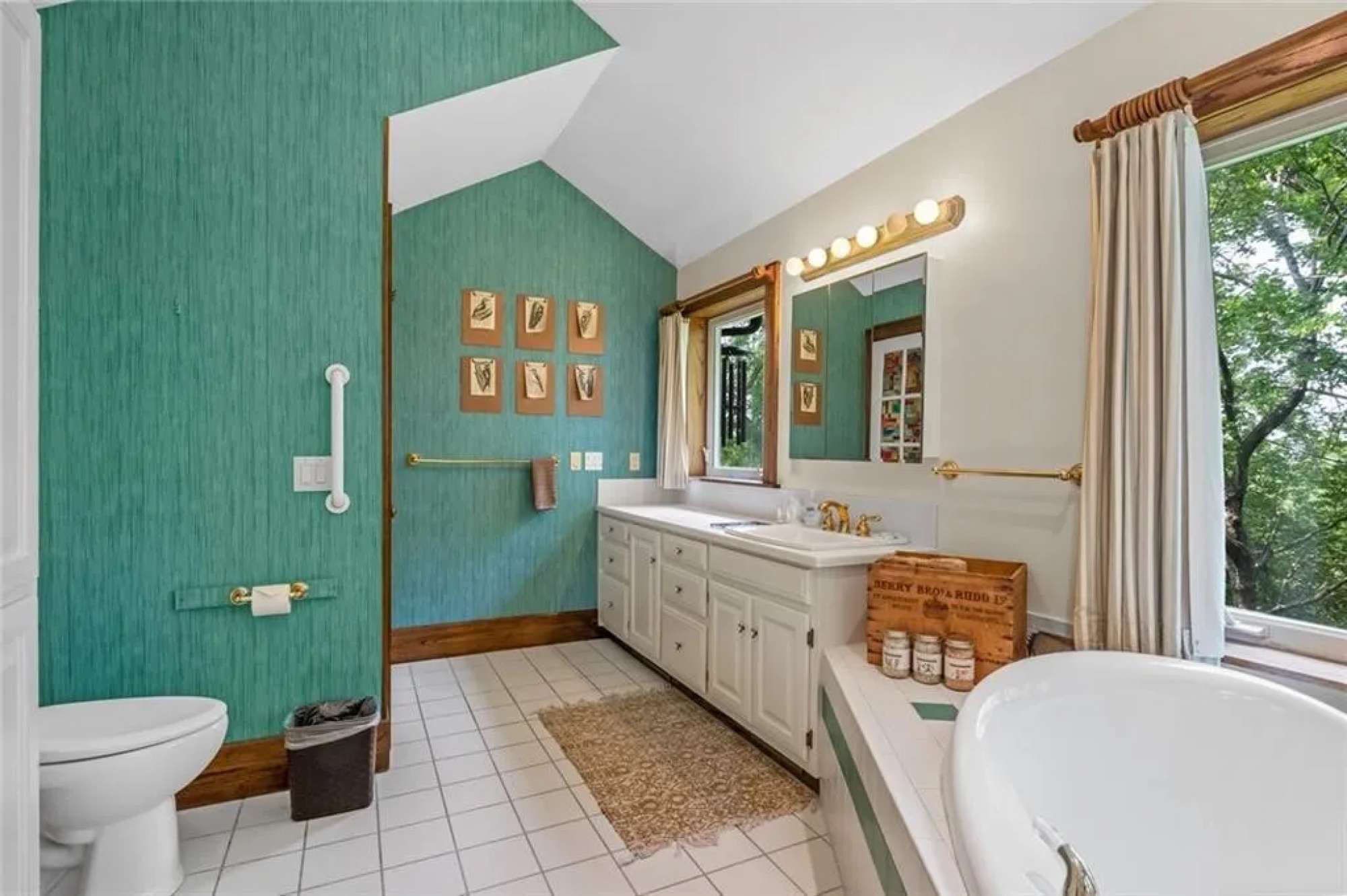 Une salle de bain avec un grand miroir, un meuble-lavabo et une baignoire. Des tableaux encadrés ornent le mur, et il y a une fenêtre avec des rideaux.