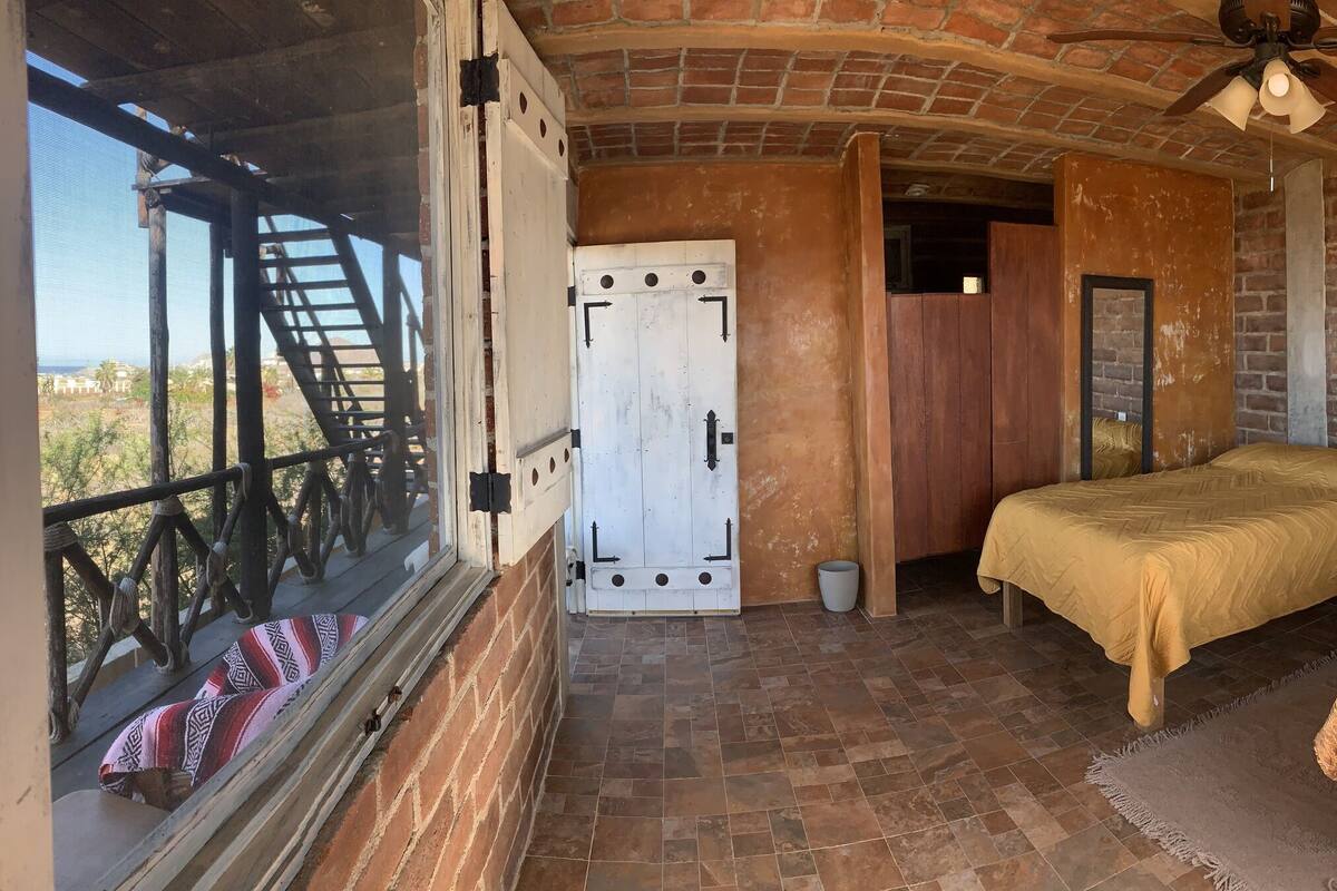 Una habitación con una cama, una mesita, un ventanal con vistas al desierto y una puerta que da a otra estancia.