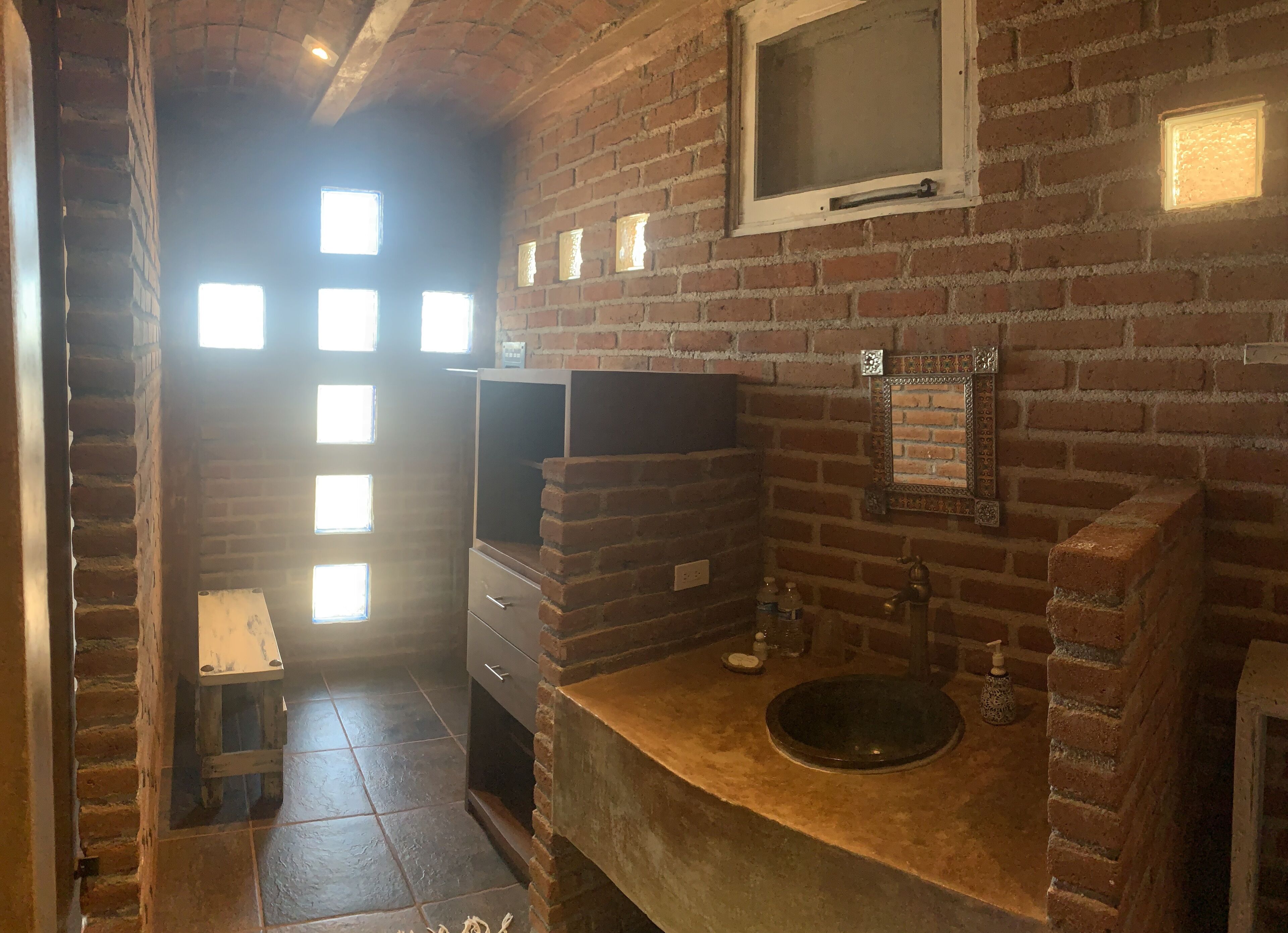 Une salle de bain rustique avec un mur en briques, un banc en bois, un lavabo avec un receveur et une fenêtre à carreaux.