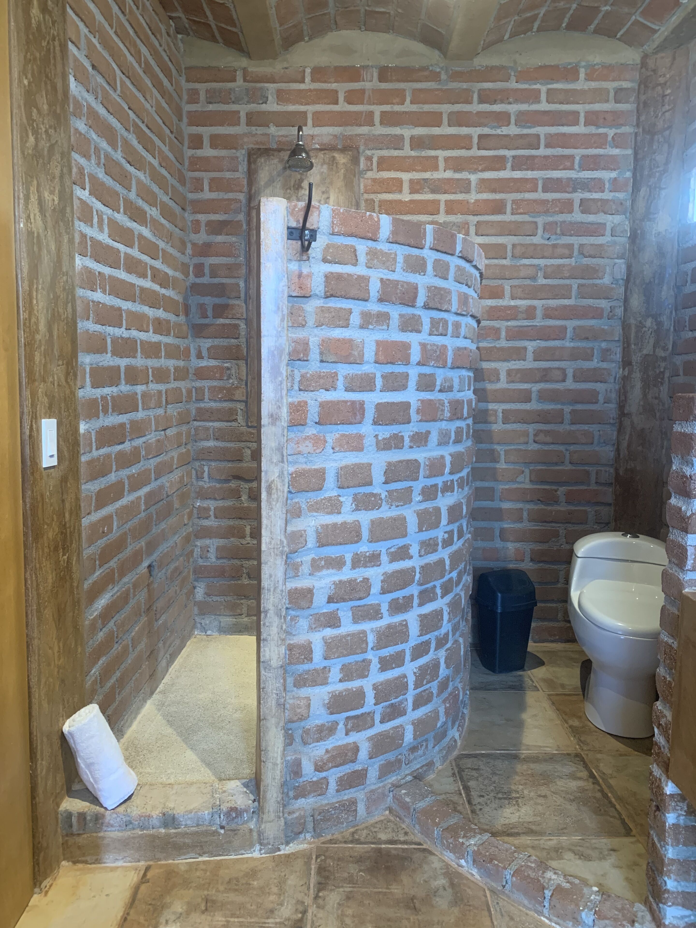 Un baño con pared de ladrillo, ducha, inodoro y cubo de basura.