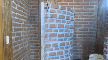 Un baño con pared de ladrillo, ducha, inodoro y cubo de basura.