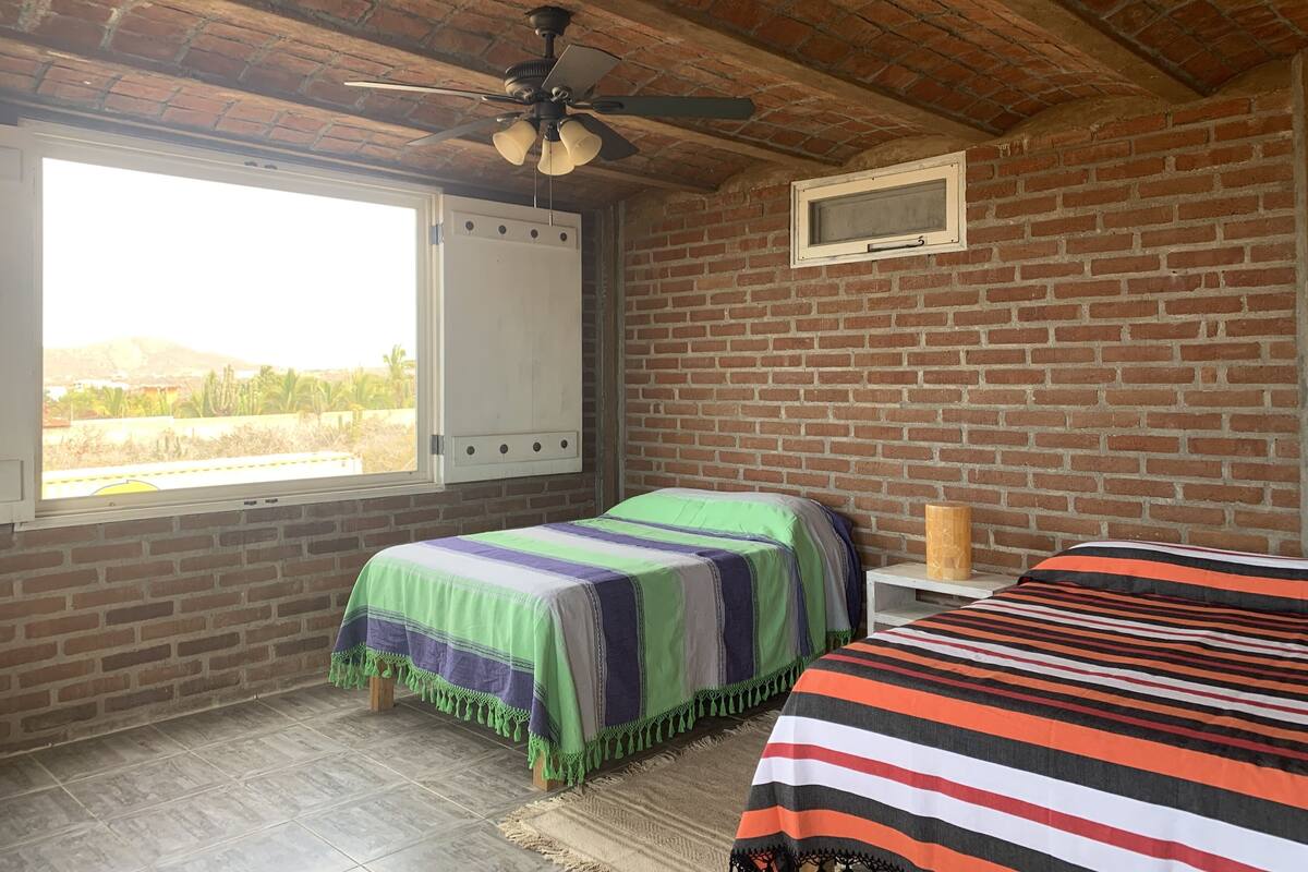 Un dormitorio con pared de ladrillo, ventana con vistas, cama con colcha a rayas y ventilador de techo.