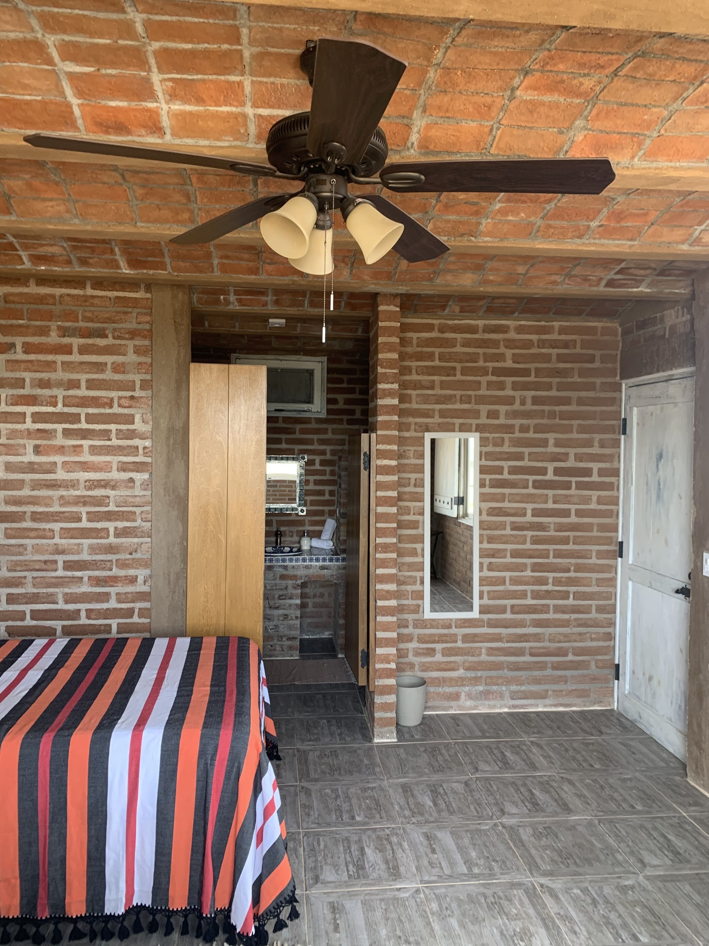 Quarto com cama, ventilador de teto, parede de tijolos, porta e um banheiro pequeno visível através de uma porta aberta.