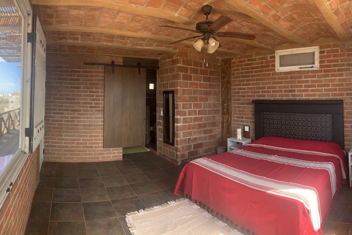 Un dormitorio con pared de ladrillo, cama con colcha roja, ventilador de techo y vistas al exterior.