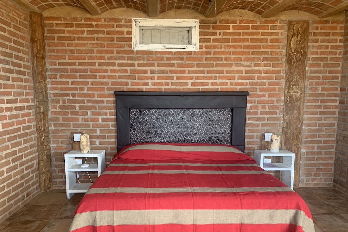 Una habitación con pared de ladrillo, una cama con manta a rayas y dos mesitas de noche con lámparas.